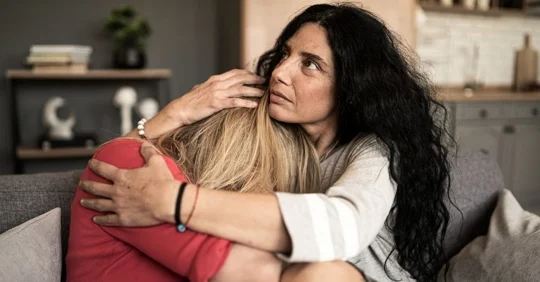 Photo of two individuals sitting on a couch, one person holding the other in a comforting manner.