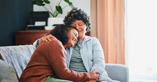 Photo of an older woman embracing a younger woman, both smiling, on a couch.