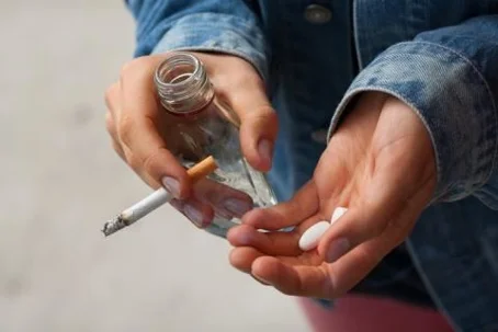 person holding a bottle of alcohol, pills, and a cigarette