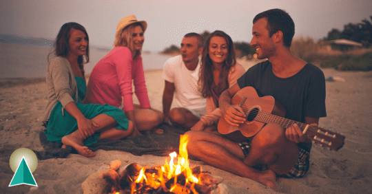 Five friends sitting on a beach around a campfire, one male playing a guitar